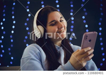 Young woman smiling with braces while listening favorite music and looking at phone against blurred blue background with lightening. 110880401
