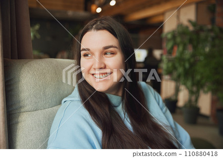 Smiling young brunette woman with braces sitting on armchair and dreamy looking at window against blurred office background. Selective focus. 110880418