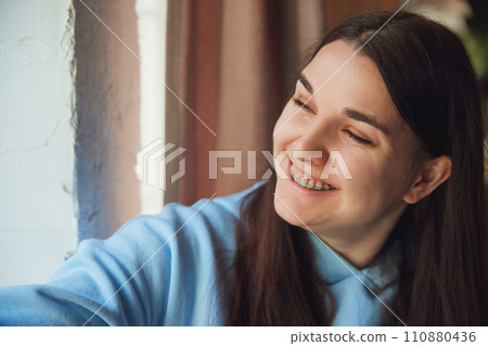 Portrait of positive young brunette woman smiling with teeth in braces sitting and thoughtfully looking at window. Selective focus. Portrait of positive young brunette woman smiling with teeth in braces sitting and thoughtfully looking at window. Selective focus. 110880436