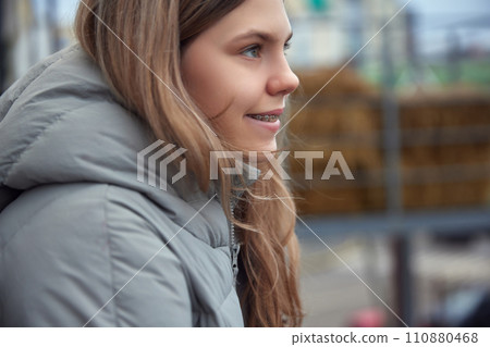 Side view portrait of youthful girl in winter coat smiling with braces, looking away against urban backdrop. Soft daylight. Side view portrait of youthful girl in winter coat smiling with braces, looking away against urban backdrop. Soft daylight. 110880468