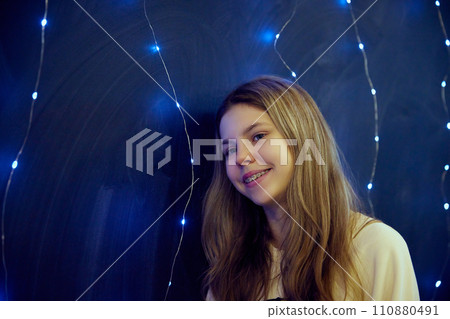 Close up photo of smiling teen girl with braces stands illuminated warm ambient lighting against blue background. Cozy indoor setting. Festive mood. Close up photo of smiling teen girl with braces stands illuminated warm ambient lighting against blue background. Cozy indoor setting. Festive mood. 110880491