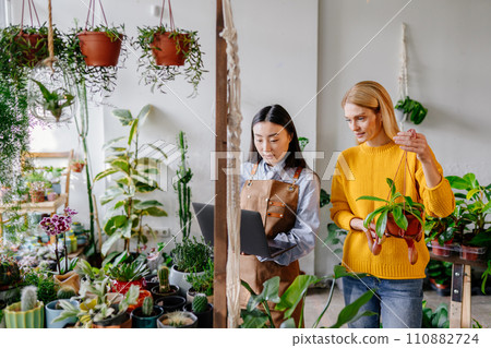 Happy middle age woman in yellow sweater in a flower shop with a purchased plant, talking with Asian female seller in apron holding laptop surround plants in plant store interior. 110882724