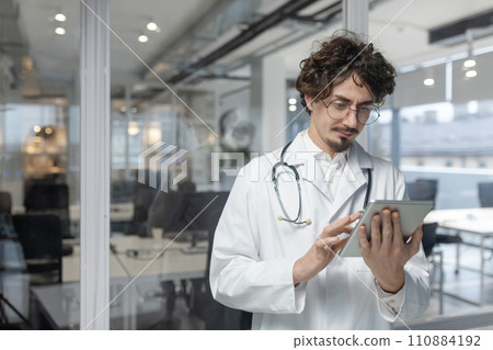 A doctor wearing a stethoscope and white medical coat looking at a tablet in a medical office. Serious reading medical reports A doctor wearing a stethoscope and white medical coat looking at a tablet in a medical office. Serious reading medical reports 110884192