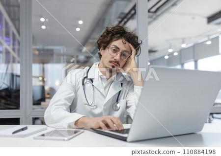A man in a white medical coat with a stethoscope sits in front of a laptop computer in a medical office. Doctor thinking using computer 110884198