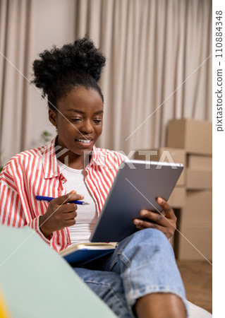 Creative woman with afro hairstyle making notes in diary while working on tablet. 110884348
