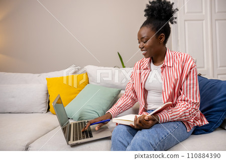 Woman with afro hairstyle writing notes in her panner and using laptop sitting on sofa. 110884390