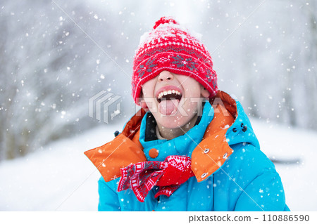 A funny little boy in blue winter clothes is walking during a snowfall. Winter outdoor activities for children. A cute child in a warm hat low above his eyes catches snowflakes with his tongue 110886590
