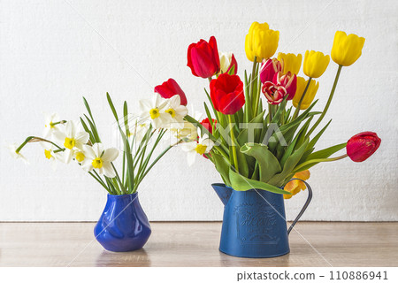 Bunch of red and yellow tulips and white daffodils in blue vases on a wooden table and white background 110886941