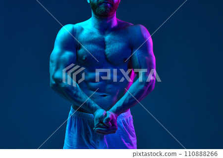 Shirtless fit male model with crossed arms posing gym shorts in neon lighting against blue studio background. 110888266