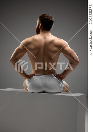 Back view portrait of muscular man, model put his hand on hips sitting on white stage in underwear against grey studio background. Back view portrait of muscular man, model put his hand on hips sitting on white stage in underwear against grey studio background. 110888278