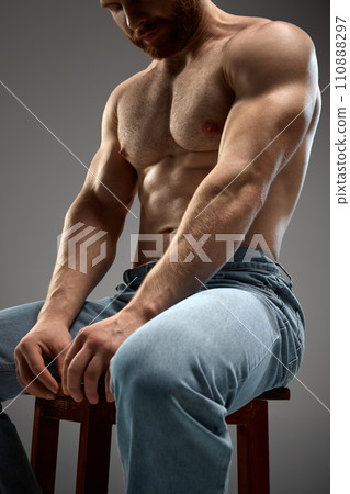 Cropped portrait of shirtless muscular man sitting on chair in denim jeans against grey background. Building muscle and strength 110888297