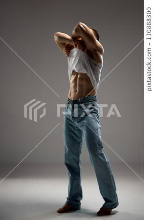 Full-length photo of man, model with an athletic and muscular body taking off his T-shirt in motion against grey studio background. 110888300