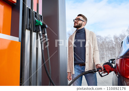 Portrait of contemporary young man stopping at gas station, looking at petrol meter and smiling while refueling car Portrait of contemporary young man stopping at gas station, looking at petrol meter and smiling while refueling car 110892176