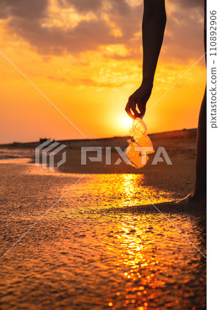 Volunteer and plastic bottle, collecting waste on the sea beach, pollution and recycling concept. hand holds a transparent plastic bottle in water lying on a sandy beach during sunset or dawn Volunteer and plastic bottle, collecting waste on the sea beach, pollution and recycling concept. hand holds a transparent plastic bottle in water lying on a sandy beach during sunset or dawn 110892906