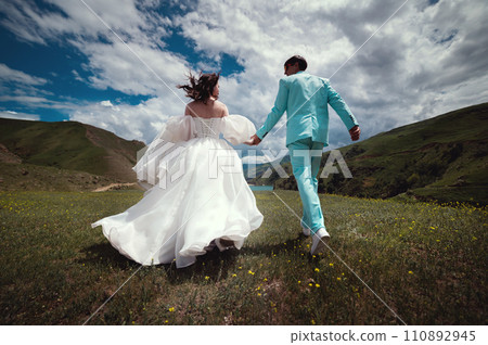 Rear view of running loving newlyweds holding hands, a bride in a wedding dress and a wife in a turquoise suit running across a field against the backdrop of green hills 110892945