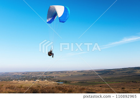A paraglider takes off from a mountainside with a blue and white canopy and the sun behind. A paraglider is a silhouette. The glider is sharp, with little wing movement. 110892965