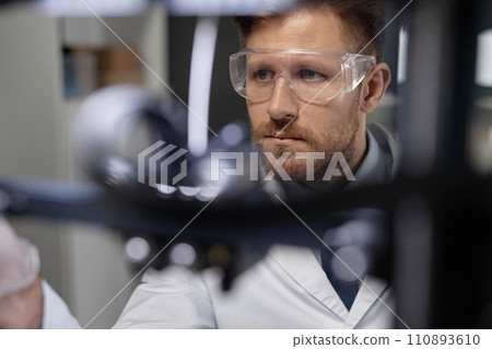 Medium close up shot of focused Caucasian male technician in protective goggles working in laboratory, look through blurred 3D printer components Medium close up shot of focused Caucasian male technician in protective goggles working in laboratory, look through blurred 3D printer components 110893610