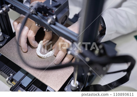 Top down view of illuminated while-colored dental model on aluminum 3D printer platform with unrecognizable technicians hands in gloves Top down view of illuminated while-colored dental model on aluminum 3D printer platform with unrecognizable technicians hands in gloves 110893611