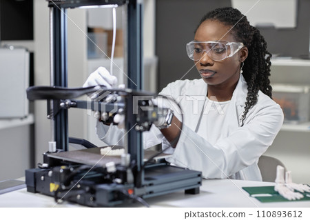 Medium close up shot of African American woman technician in protective glasses adjusting filament strap of 3D printer while sitting in laboratory Medium close up shot of African American woman technician in protective glasses adjusting filament strap of 3D printer while sitting in laboratory 110893612