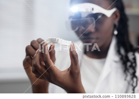Close up shot of white-colored 3D toothed jaw bone sample in hand illuminated by magnifying glasses light on face of African American female technician 110893628