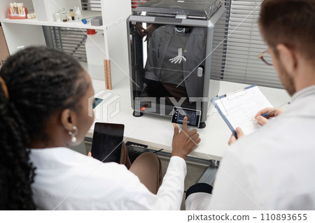 High angle shot with back view of female engineer selecting 3D printer settings on user interface and holding tablet while male coworker filling in registration card, both sitting at laboratory table High angle shot with back view of female engineer selecting 3D printer settings on user interface and holding tablet while male coworker filling in registration card, both sitting at laboratory table 110893655