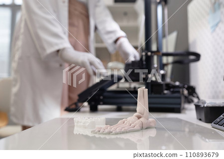 Close up shot of produced white-colored 3D dental and bone samples on acrylic print bed in laboratory, unrecognizable technician working in blurred background 110893679