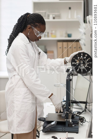 Vertical shot of Black woman engineer in lab coat and protective glasses setting up 3D printer for work in laboratory Vertical shot of Black woman engineer in lab coat and protective glasses setting up 3D printer for work in laboratory 110893681