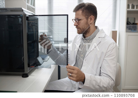 Medium long shot of Caucasian man engineer in lab coat and eyeglasses taking produced bone sample out of enclosed 3D printer in laboratory 110893708