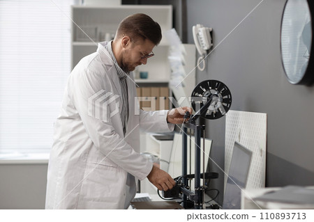Caucasian male engineer in lab coat inserting white-colored filament strand into extruder of 3D printer in laboratory 110893713