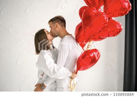 A girl kisses a guy on Valentine's Day holding a bunch of red foil balloons in the shape of a heart 110893884