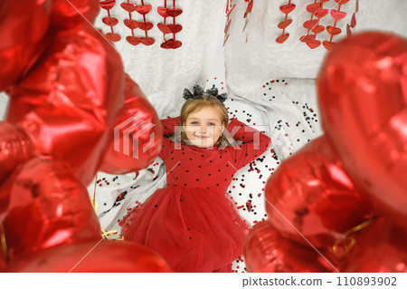 A little girl in a red polka dot dress, surrounded by heart-shaped balloons, lies on the floor looking at the camera. 110893902