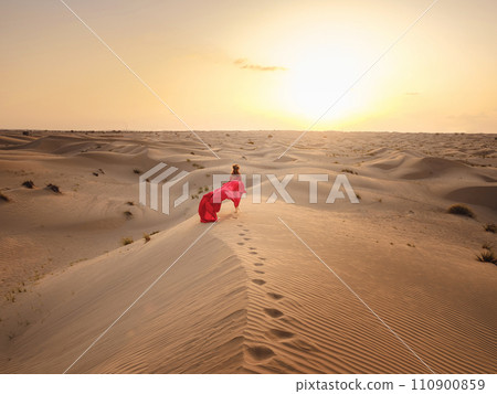 Desert adventure. Young arabian Woman in red silk dress in sands dunes of UAE desert at sunset, fantastic view. The Dubai Desert Conservation Reserve, United Arab Emirates. Desert adventure. Young arabian Woman in red silk dress in sands dunes of UAE desert at sunset, fantastic view. The Dubai Desert Conservation Reserve, United Arab Emirates. 110900859