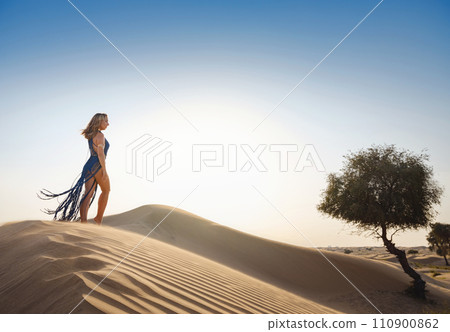 Desert adventure. Young arabian Woman in sexy boho blue makrame dress in sands dunes of UAE desert at sunset. The Dubai Desert Conservation Reserve, United Arab Emirates. Desert adventure. Young arabian Woman in sexy boho blue makrame dress in sands dunes of UAE desert at sunset. The Dubai Desert Conservation Reserve, United Arab Emirates. 110900862