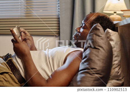 Mature black man in homewear lying on bed and looking through old faded photographs reminiscing past Mature black man in homewear lying on bed and looking through old faded photographs reminiscing past 110902513