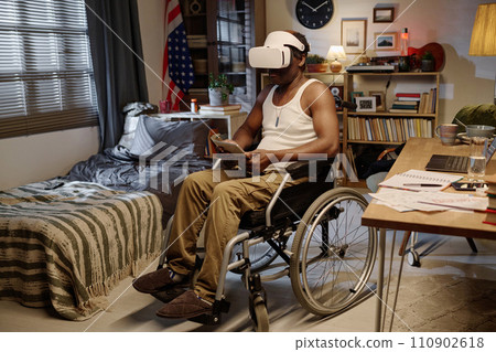Black man in wheelchair wearing virtual reality headset and holding digital tablet sitting at home Black man in wheelchair wearing virtual reality headset and holding digital tablet sitting at home 110902618