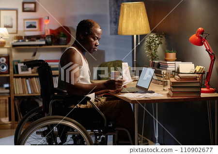 Mature african american man in wheelchair sitting at desk in his bedroom and looking through bills 110902677
