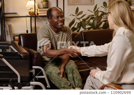 Joyful black man with disability shaking hands with female psychologist sitting on sofa at office 110902699