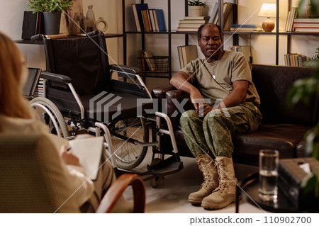 Black veteran with disability sitting on couch in relaxed pose and talking to therapist taking notes 110902700