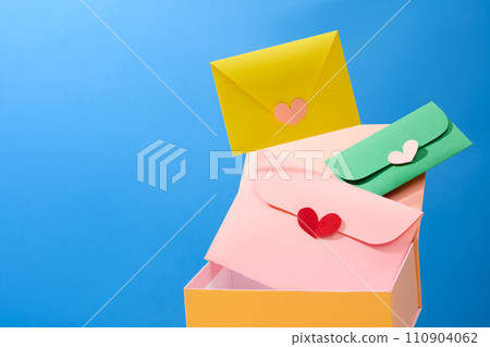Front view of a yellow gift box with cute handmade envelopes are flying on blue background. Creative ideas for advertising on International Women Day. Copy space Front view of a yellow gift box with cute handmade envelopes are flying on blue background. Creative ideas for advertising on International Women Day. Copy space 110904062