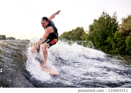 Young athletic man energetically riding wakeboard. Handsome caucasian guy glides on the wave like a real surfer. Active lifestyle and extreme sports. Summertime watersports activity. 110904351