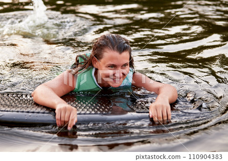 Young beautiful sporty girl in a green life jacket swims in the water with a wakeboard in her hands. Happy sportswoman is preparing for a steep wakeboard ride. Active lifestyle, healthy hobby. 110904383