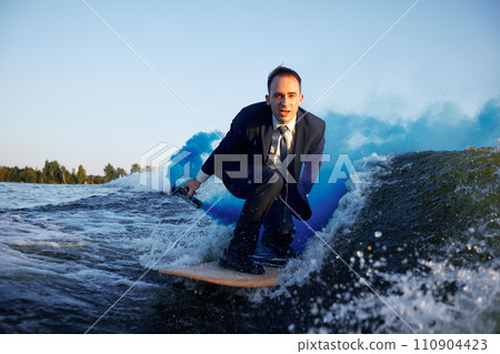 Young man in classic suit with blue smoke bomb in his hand rides wakeboard on a lake near city. Happy clerk escaped from stuffy office to take up active sport. Best summer leisure after routine work. 110904423