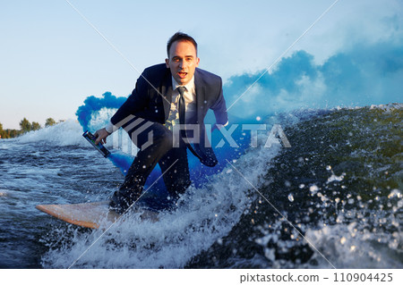 Young man in classic suit with blue smoke bomb in his hand rides wakeboard on a lake near city. Happy clerk escaped from stuffy office to take up active sport. Best summer leisure after routine work. Young man in classic suit with blue smoke bomb in his hand rides wakeboard on a lake near city. Happy clerk escaped from stuffy office to take up active sport. Best summer leisure after routine work. 110904425