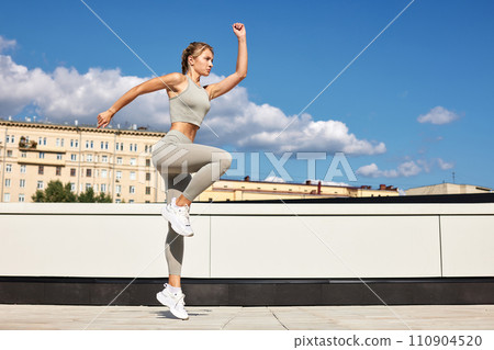 Fit woman exercising outdoors. Healthy young sportswoman doing fitness. An athlete makes jumps, does functional training on the street on a bright sunny day, smiles contentedly, in a sports suit Fit woman exercising outdoors. Healthy young sportswoman doing fitness. An athlete makes jumps, does functional training on the street on a bright sunny day, smiles contentedly, in a sports suit 110904520
