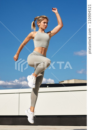 Fit woman exercising outdoors. Healthy young sportswoman doing fitness. An athlete makes jumps, does functional training on the street on a bright sunny day, smiles contentedly, in a sports suit Fit woman exercising outdoors. Healthy young sportswoman doing fitness. An athlete makes jumps, does functional training on the street on a bright sunny day, smiles contentedly, in a sports suit 110904521