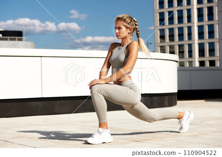 Middle-aged athlete enjoys an evening workout at sunset, stretches her legs, does lunges, squats before jogging. Sportswoman training outdoors, in an urban environment Middle-aged athlete enjoys an evening workout at sunset, stretches her legs, does lunges, squats before jogging. Sportswoman training outdoors, in an urban environment 110904522
