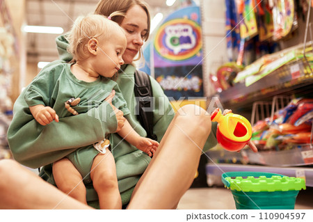 Young attractive woman with her cute baby sitting on the floor in toy store. Beautiful mom and her little blonde daughter shopping in the mall and having fun. Family weekend, happy childhood. 110904597
