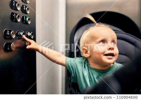 Tiny cute baby sitting in a baby stroller presses the floor button in the elevator. A little blonde in a carriage goes up or down the elevator with her mom. Parental control. 110904600