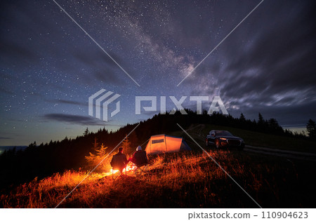 Happy couple staring at starry sky with clouds. Young people sitting in chairs and warming by campfire near tourist tent and off-road car. Travelling couple having a rest together. Night camping. 110904623