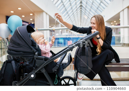 Young pretty woman holding carriage with her child. Happy mother and cute little blonde girl in pink clothes play, laugh and have fun. Mom with a baby in a stroller walking around shopping center. Young pretty woman holding carriage with her child. Happy mother and cute little blonde girl in pink clothes play, laugh and have fun. Mom with a baby in a stroller walking around shopping center. 110904660